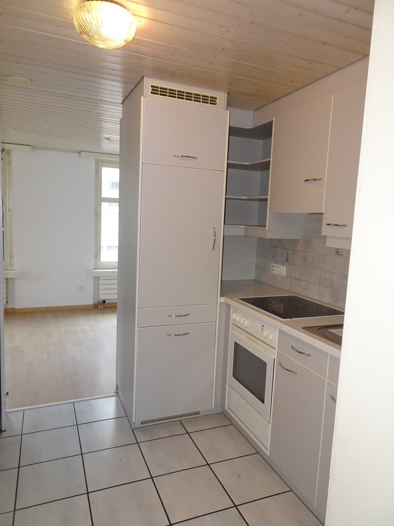 The image shows a small kitchen with white cabinets, a built-in refrigerator, and a stove. The floor is tiled, and there is a ceiling light fixture. The kitchen appears to be part of a larger living space.