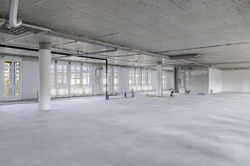 Unfinished office space with exposed concrete, large windows, and unfinished fixtures.