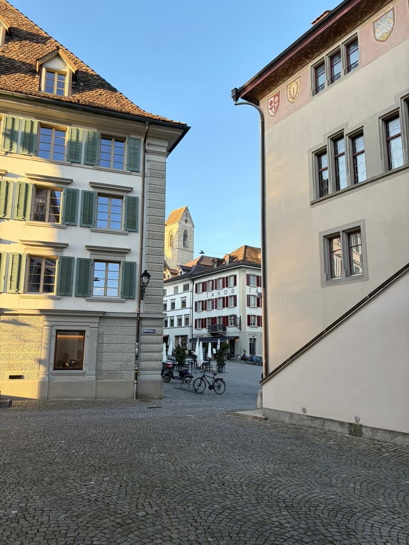 Two buildings with multiple windows, a cobblestone street, bicycles parked, and a church tower in the background.