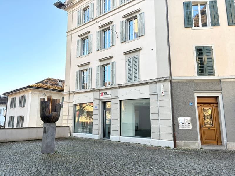 A three-story building with a modern architectural design. The building has a mix of white and beige walls, with several windows and shutters. The entrance is marked by a wooden door and a signboard. The building is situated on a cobblestone street, with