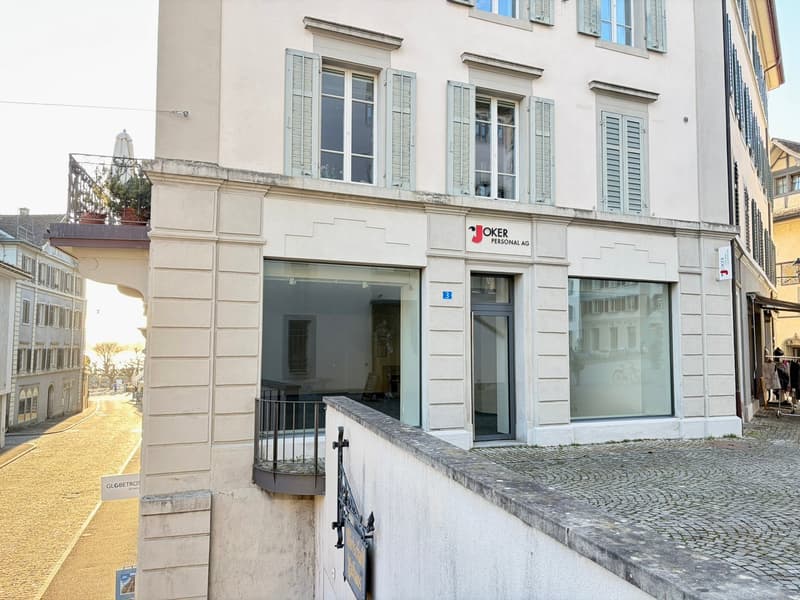 White building with glass windows, balcony with metal railing, entrance with glass door, signboard with text 'Joker Personal AG', cobblestone pavement