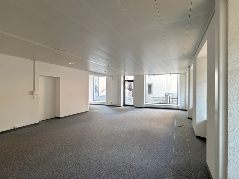 Empty room with white walls, gray carpet, ceiling lights, and glass doors.
