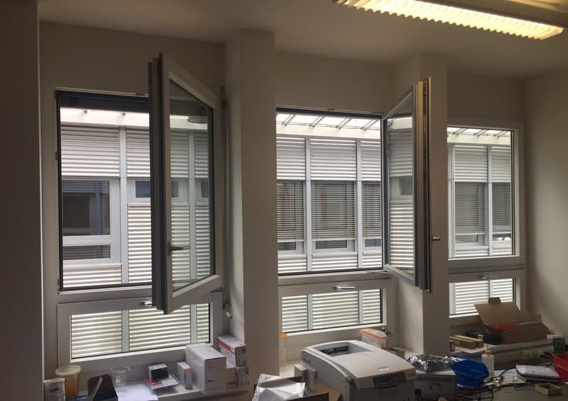 The image shows an office space with large windows, blinds, and various office equipment and supplies such as a printer, boxes, and other items. The space appears to have a balcony or terrace visible through the windows.