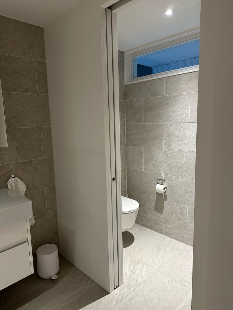 modern bathroom with white tiles, white toilet, white sink, small window, trash can
