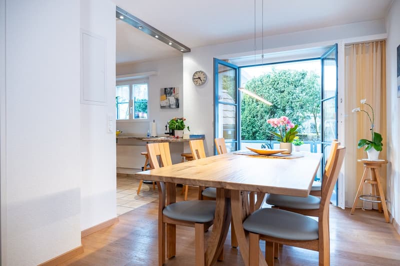 Dining room with wooden table, chairs, plants, open door to balcony, kitchen in background