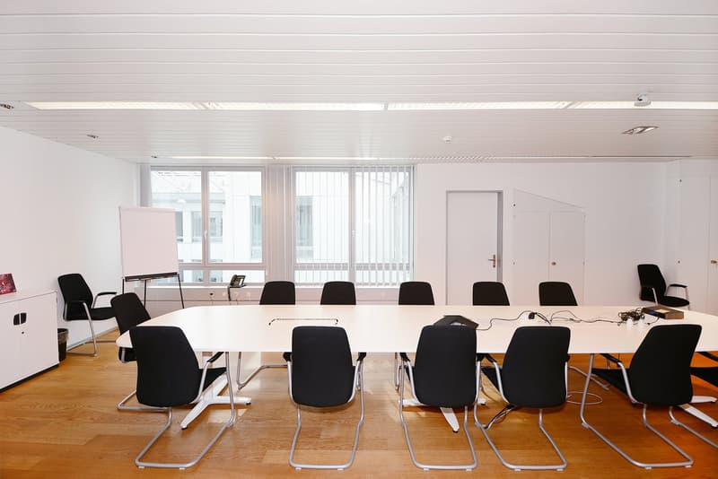 A modern, rectangular meeting room with white walls and wooden floors. The room features a large white table surrounded by black chairs. There is a whiteboard on an easel in the corner, and a whiteboard is mounted on the wall. There are windows with blind