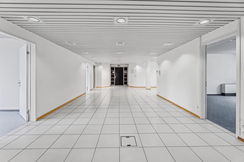 Long, wide hallway with white walls, a tiled floor, and a suspended ceiling with recessed lighting. The hallway appears to be in a commercial or office building, with doors leading to other rooms or offices on either side.