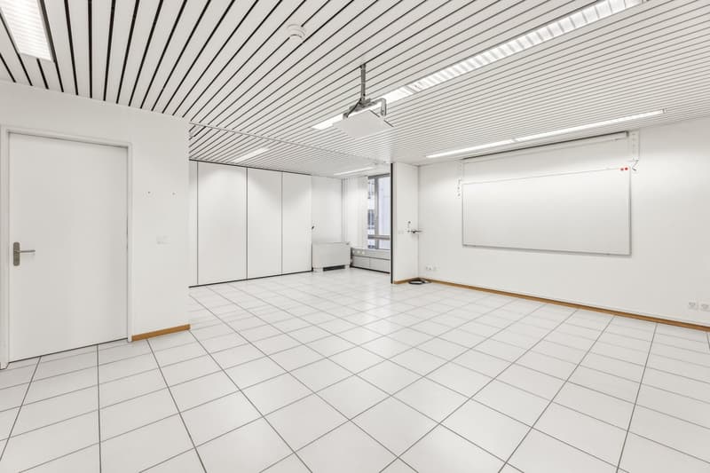 Large open room with white walls, tiled flooring, and a suspended ceiling with linear lighting. The room has a whiteboard on one wall and built-in storage cabinets along another wall.