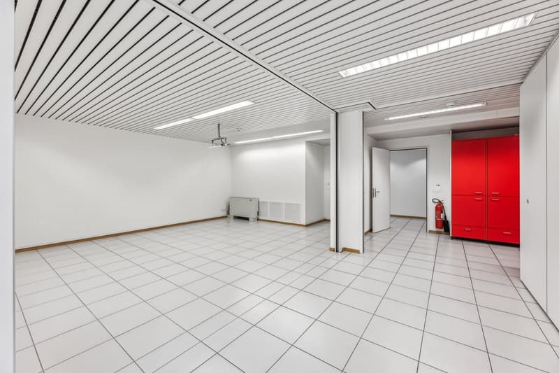 Large open office space with white walls, a tiled floor, and a suspended ceiling with linear lighting. The room has a storage unit and a fire extinguisher visible.