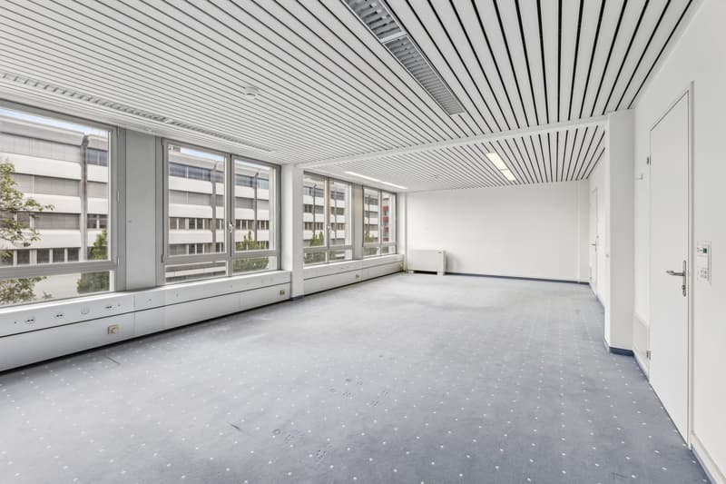 Large open office space with high ceilings, large windows, and a white tiled floor. The room has a minimalist, modern design with a white ceiling and walls.