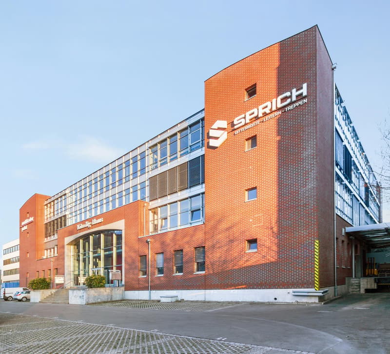 Exterior view of a modern office building with red brick walls and large glass windows. A clear blue sky is visible. The main feature is a large white and grey logo reading 'SPRICHS' on the front wall. The entrance has automatic glass doors and stairs. A