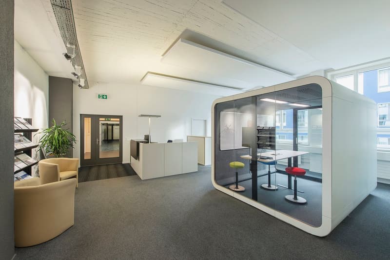 Contemporary design, white furniture, gray carpet, glass-enclosed cubicle, two stools, desk, modern lighting fixtures, potted plant, exit sign, two chairs