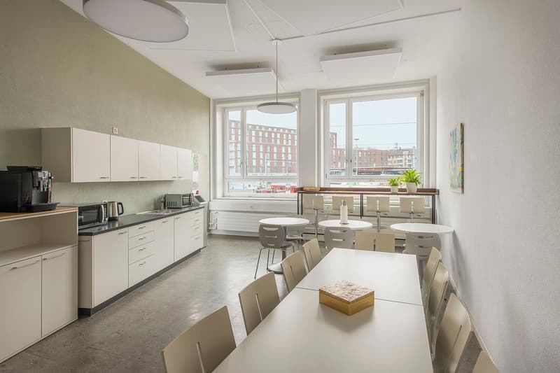 White kitchen cabinets, microwave, coffee machine, several tables and chairs, large windows