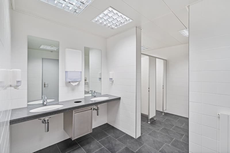 a bathroom with two sinks, faucets, mirrors, paper towel dispensers, and a grey tile floor