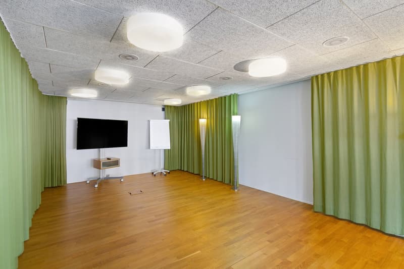 An empty room with wooden flooring, white walls, and two green curtains. A television is mounted on the wall, with a whiteboard nearby, and two tall lamps on the side. The ceiling is lit with several circular lights.