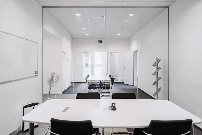 Conference room with white walls, glass partition, white table, chairs, whiteboard, floor fan, and metal rack
