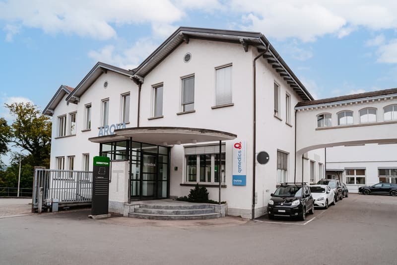 Large white building, several windows, arched entrance, cars parked in front, green sign