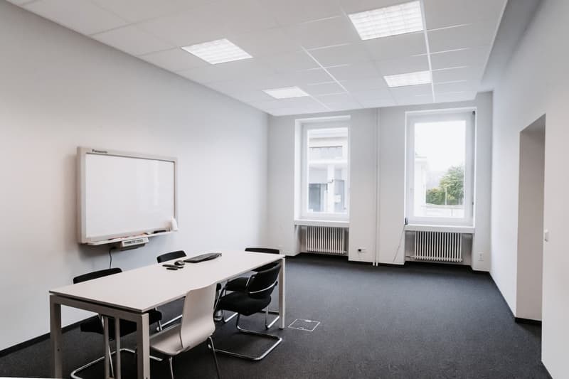 Empty room with white walls, black carpet, long white table, white chairs, white board, two windows with white radiators