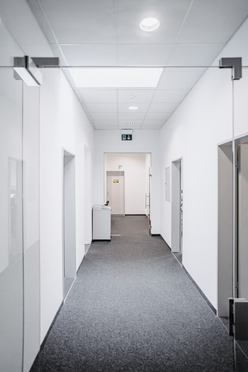 Empty hallway, carpeted floor, ceiling lights, glass doors, emergency exit sign, white walls, white ceiling