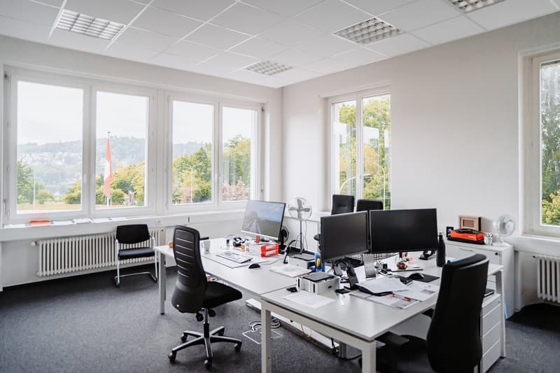 Spacious, contemporary office space with large windows providing natural light and scenic views, well-lit with multiple computers, office chairs, and a central desk.