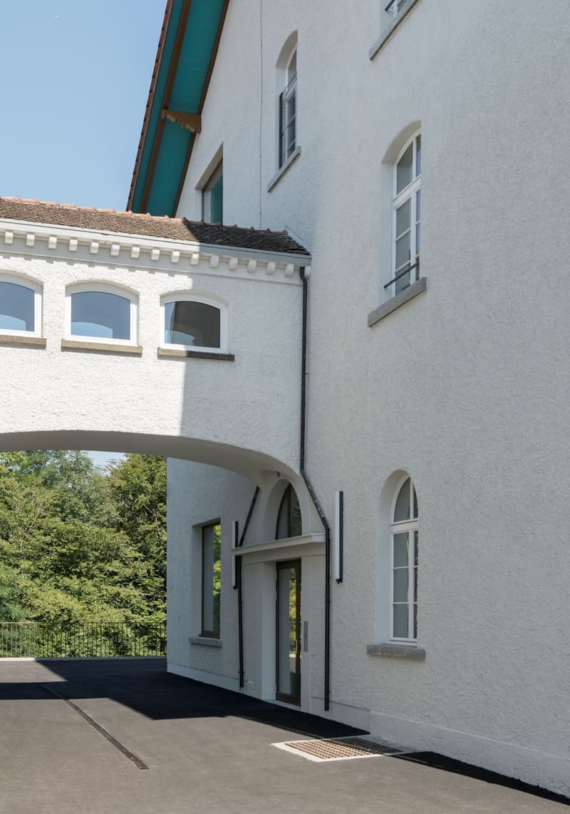 A modern white house with multiple windows, an arched entryway, a fence, and a paved car park in front.