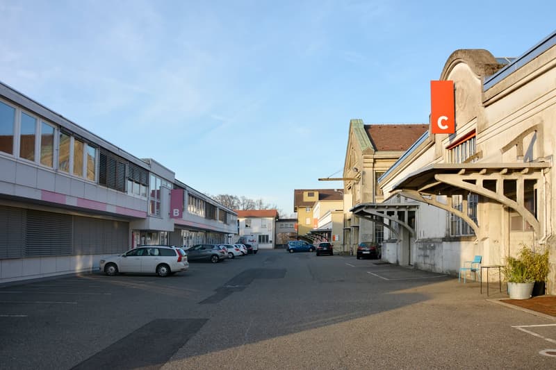 The image shows a commercial area with several buildings, including a large building with a red 'C' sign on it. There are parked cars in the paved parking lot in the foreground, and the buildings have a mix of modern and older architectural styles.