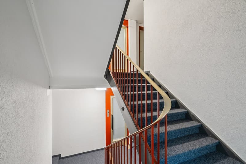 Spiral staircase with blue carpet and red handrail, white walls, orange door with glass panels, lamp on wall, small window