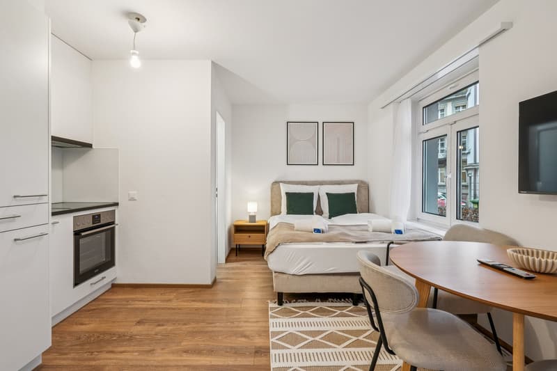 small kitchen area with oven, bed with pillows and duvet, bedside table with lamp, small dining table with chairs, open window