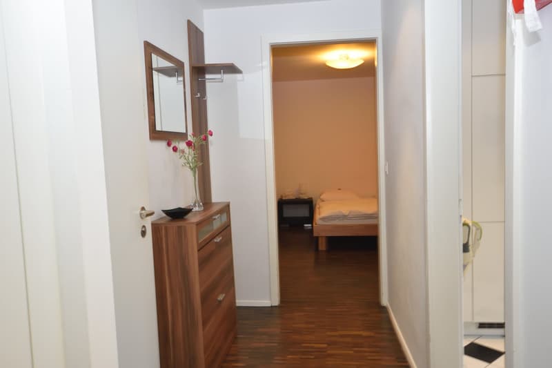 Interior hallway with white walls, wooden floor, wooden cabinet, flowers, mirror and single bed in adjacent room.