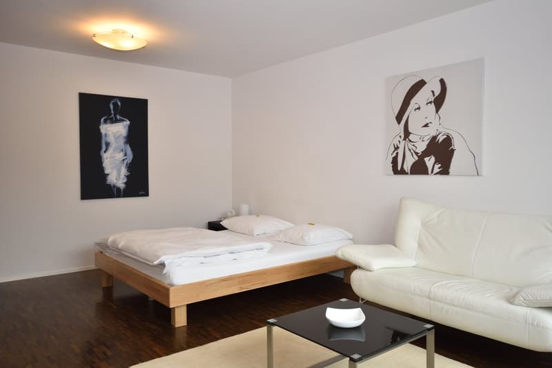 White walls, wooden floor, white couch, double bed, black coffee table, artworks
