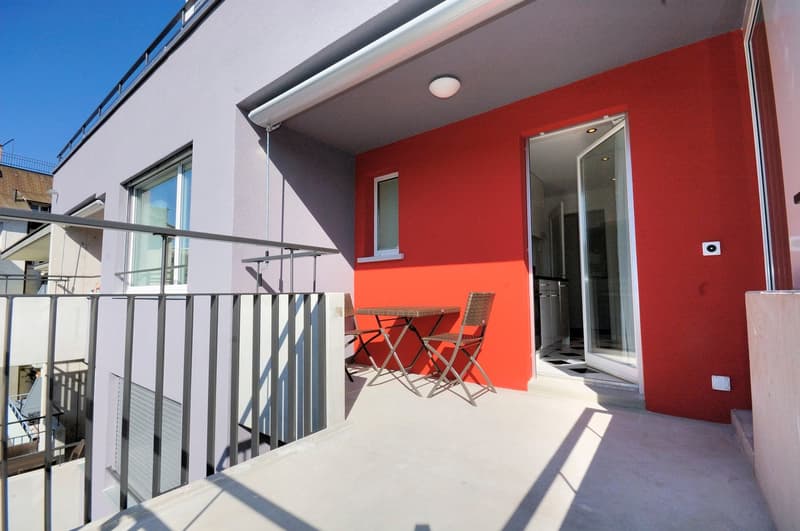 Outdoor balcony with table and chairs, metal railings, red wall, entrance door, white ceiling lamp