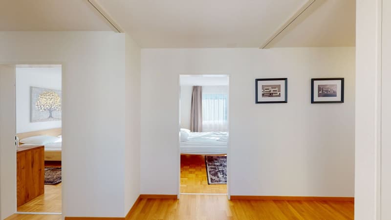 A hallway leading to a bedroom. The hallway features white walls, wooden flooring, and a few decorative elements.