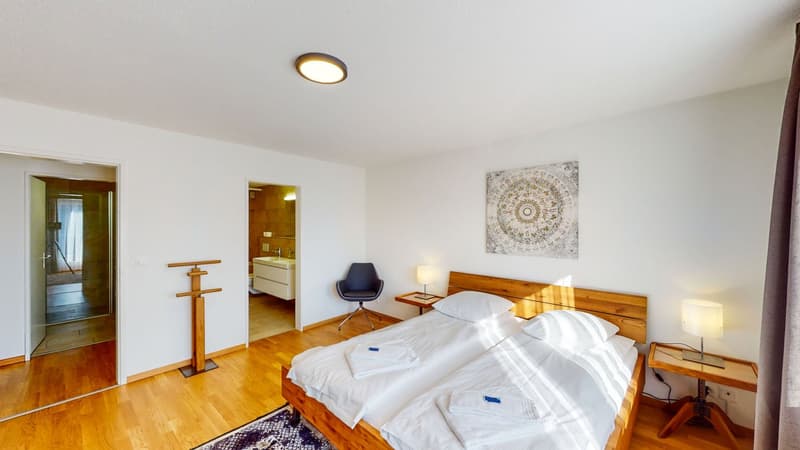 Two single beds, two nightstands with lamps, blue chair, coat rack, artwork on the wall, bathroom doorway, bathroom sink, and shower