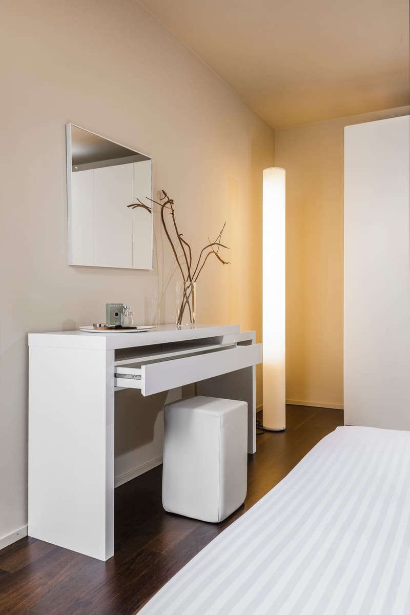 minimalist bedroom with white desk, white stool, white bed, mirror, lamp, vase, and plant on the desk