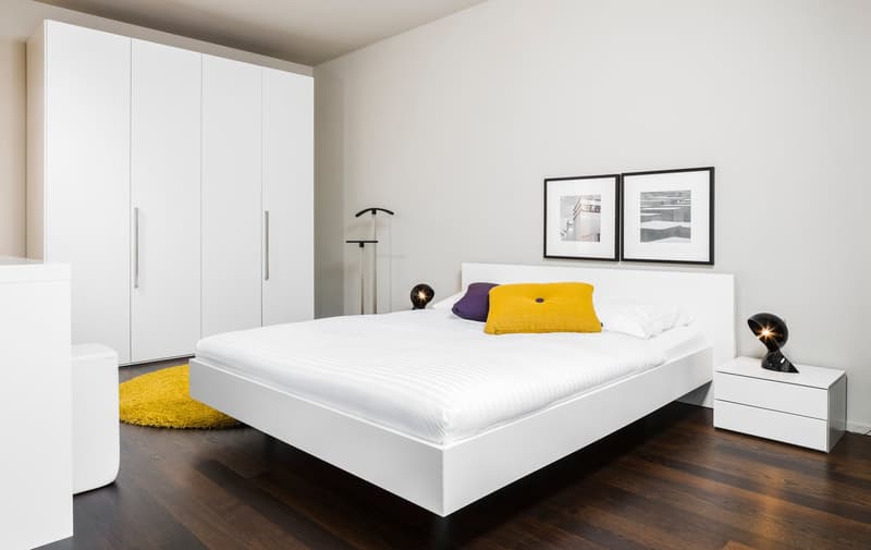 A modern, minimalist bedroom with white walls, wooden floor, and a white bed frame. The bed has white sheets and two pillows. A yellow carpet and a white nightstand with a lamp are placed on the side. There are two picture frames on the wall. A white ward