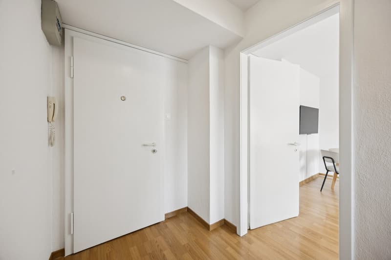 An empty room with white walls, wooden floor, two white doors, a phone mounted on the wall, and a television on the wall of the next room.