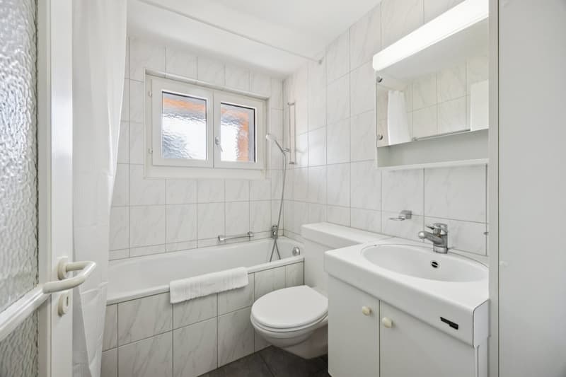 Bathroom with white tiled walls, bathtub, toilet, sink, mirror, showerhead, and towel rack