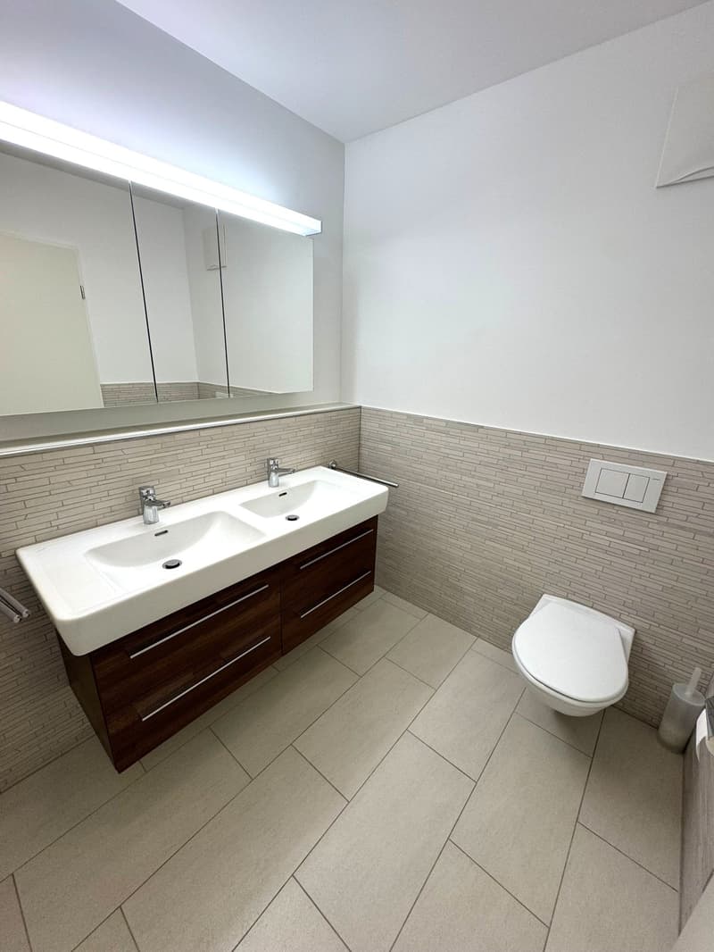 modern bathroom with double sink, large mirror, white toilet, tiled floor, wall-mounted light