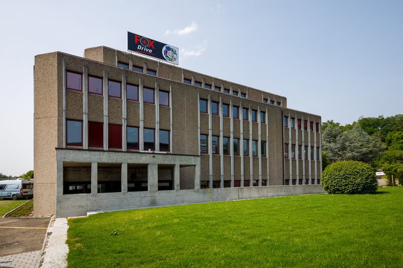 Multi-story concrete and glass office building with a Fox Drive logo on the top. The building has a rectangular shape with rows of windows and a grassy area in front.