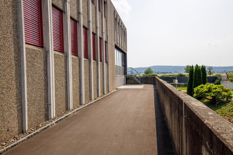 A large concrete building with red shutters, surrounded by a paved walkway and landscaped gardens. The building appears to have a balcony or terrace overlooking the scenic outdoor area.