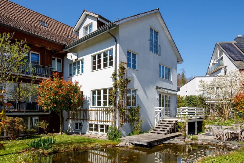2 story house, white exterior, brown roof, large windows, wooden deck, small pond, garden