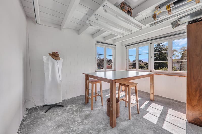 This image shows a bright and airy studio or workspace with large windows providing a scenic view of the outdoors. The room has a high, vaulted ceiling with exposed white beams. There is a wooden table with stools, and a hat hanging on the wall. The floor