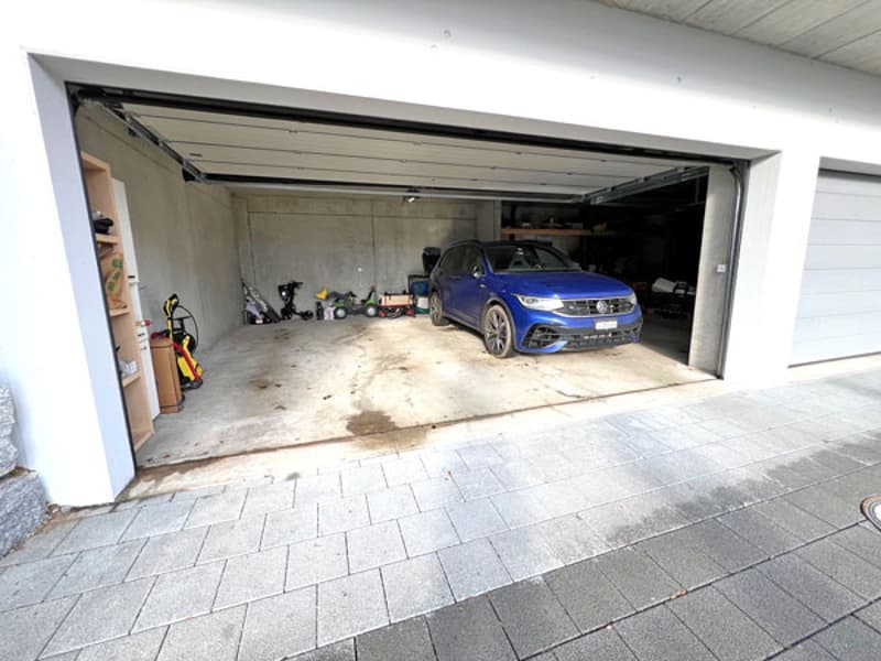 Ample garage space with a concrete floor, side shelves, and a blue car parked inside. Two garage doors, one open and one closed.