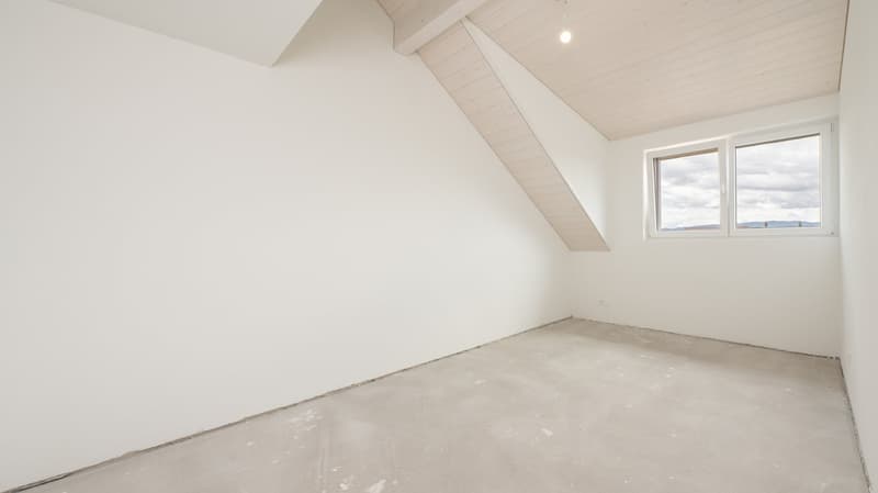 This image shows an empty room with white walls, a slanted ceiling, and a large window. The room appears to be unfinished, with a concrete floor and no furniture or decor present.