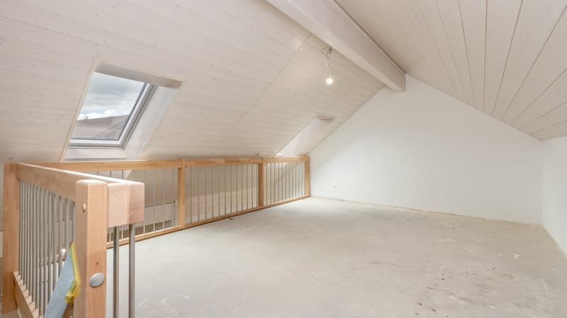 Spacious attic room with wooden beams, white walls, and a skylight window. The room has an open floor plan with a wooden railing along the upper level.