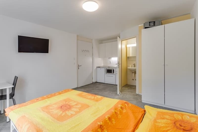 Bedroom with bed, desk, chair, TV, microwave, sink, cabinets, and a door to the bathroom.