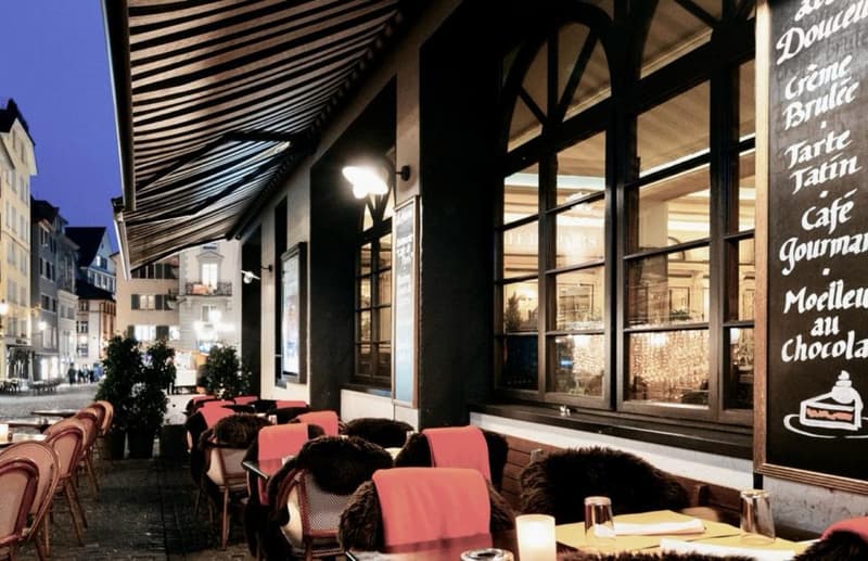 exterior of a French cafe with awning, tables, chairs, large windows, outdoor light, potted plants, signboard