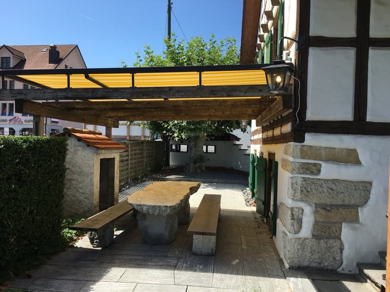 Outdoor dining table with benches under a pergola, stone patio, lamp