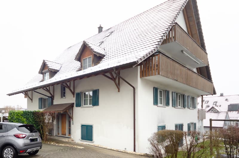 White 2-story house, wooden balcony, brown roof, green shutters, driveway