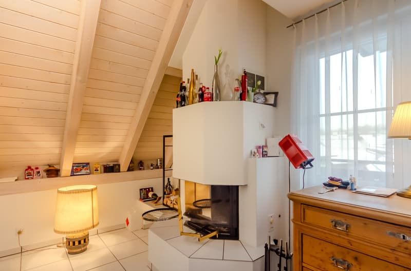 Attic room, sloped ceiling, fireplace, white tile flooring, wooden dresser, desk with lamp, glass door